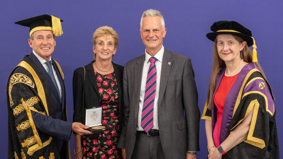 Four people stand against a purple background; two in academic regalia, one presenting a medal, and two in formal attire.